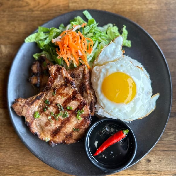 Vietnamese grilled pork chop with a fried egg, rice, salad, and chili on a black plate.