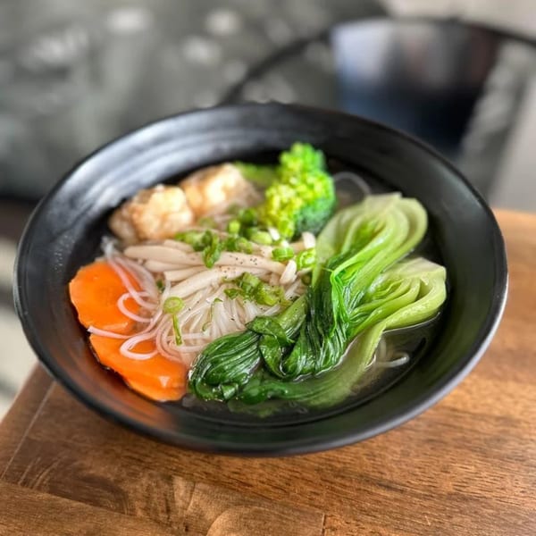 A black bowl of Vietnamese pho with noodles, bok choy, carrots, broccoli, and meatballs.