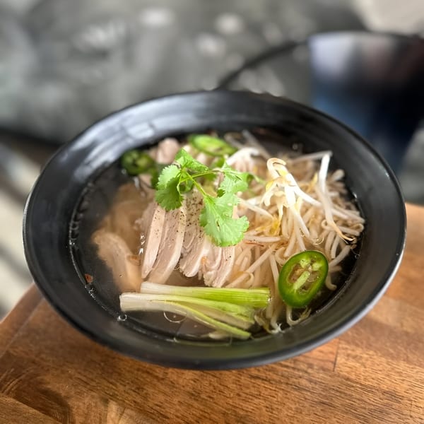 A bowl of Pho Tám Cali chicken pho with bean sprouts, jalapeños, and cilantro on a wooden table.
